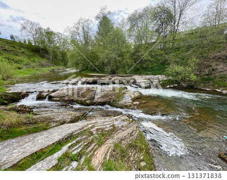 Picturesque stream flows over flat, layered rocks, forming gentle cascades in lush, green landscape. Trees and shrubs adding to natural beauty of scene. Clear water reflects serene surroundings. Picturesque stream flows over flat, layered rocks, forming gentle cascades in lush, green landscape. Trees and shrubs adding to natural beauty of scene. Clear water reflects serene surroundings. 131371838