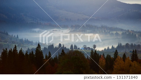 Misty autumn morning in mountain valley. Layers of hills bathed in soft light, with patches of sunlight breaking through fog, illuminating autumn foliage. Interplay of light and shadow. 131371840