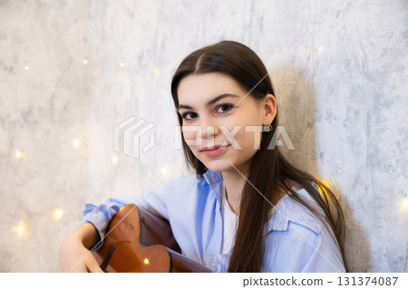 A young woman sits against a textured wall, strumming her guitar and smiling softly. The warm atmosphere is enhanced by twinkling lights in the background, creating a peaceful vibe. 131374087