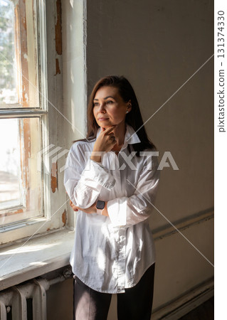 Woman in a white shirt standing thoughtfully near a window in a bright room during daytime. A business portrait of woman in creative profession. Architect, designer, artist, photographer, doctor 131374330