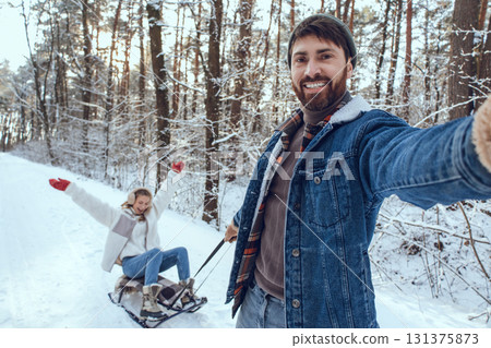 Bearded young man pulling sled with his wife sitting on it Bearded young man pulling sled with his wife sitting on it 131375873