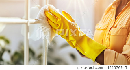 Cleaning window with yellow gloves and cloth, sunlight streaming in background, spring cleaning concept, 131376745