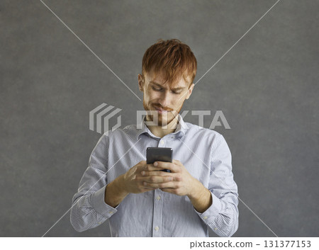 Portrait of smiling young man using mobile phone standing over grey studio wall 131377153