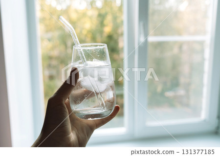 Hand holding a glass of iced water with a reusable glass straw by a bright home window healthy hydration lifestyle 131377185