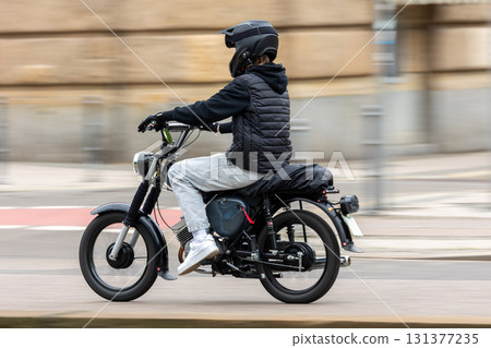 Young motorcyclist in casual clothing and helmet rides black motorbike along urban street. Dynamic shot with motion blur emphasizes speed, energy and the urban lifestyle of two-wheeled transport 131377235