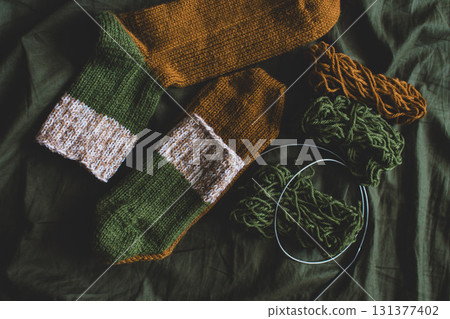 Cozy knitted socks with yarn ball and knitting needles. Striped wool socks on dark green sheet. Cozy autumn colours. Woven crochet close up. Knitting hobby. Knitwear concept. Needlework background.  131377402