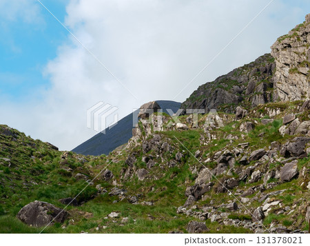 This showcases the rocky terrain and lush greenery of the Gap of Dunloe in Ireland. A mountain rises in the background under a partly cloudy sky. This showcases the rocky terrain and lush greenery of the Gap of Dunloe in Ireland. A mountain rises in the background under a partly cloudy sky. 131378021