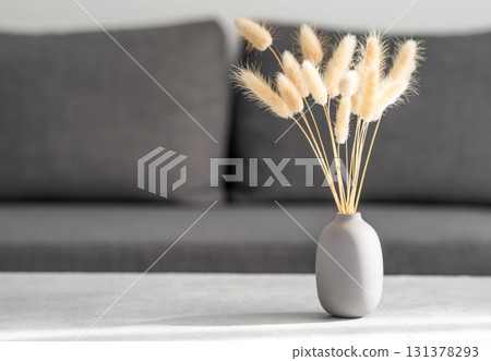 Dried grass "rabbit tail" in a matte vase standing on a concrete table against a soft gray sofa. 131378293
