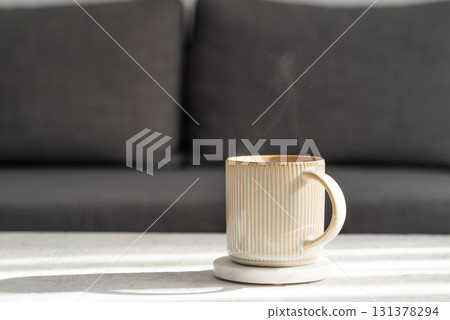 Ceramic cup of coffee on light table with morning shadows, in front of a gray sofa. 131378294