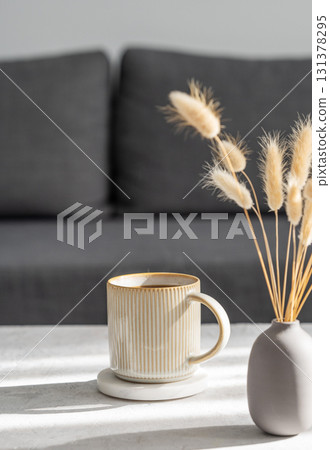Beige ceramic cup of coffee with steam on a light table with soft shadows, vase and notebook Beige ceramic cup of coffee with steam on a light table with soft shadows, vase and notebook 131378295