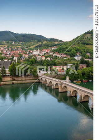 An old stone bridge with arches spans the tranquil Drina River, surrounded by green hills 131378313