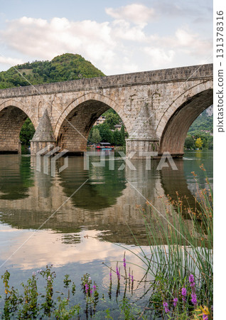 An ancient stone bridge with arches spanning the tranquil Drina River with wild flowers  131378316