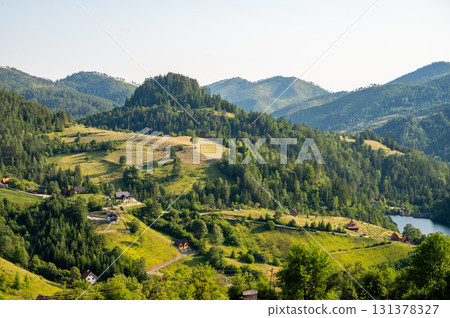 Lush green valley with rural houses surrounded by forested mountains under clear blue sky. 131378327