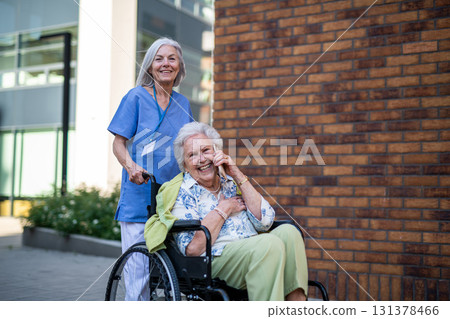 Older nurse pushing senior woman in wheelchair while making phone call. 131378466