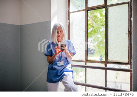 Tired nurse taking break, standing by old widnow and texting. 131378476