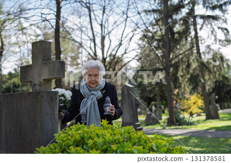 Elderly widow bringing flowers to a grave at cemetery. Elderly widow bringing flowers to a grave at cemetery. 131378581