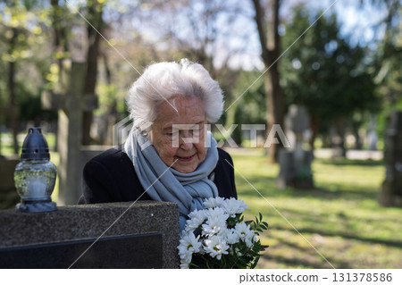 Elderly widow bringing flowers to a grave at cemetery. Elderly widow bringing flowers to a grave at cemetery. 131378586