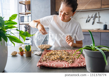 Young woman pouring stones into a glass container while preparing a terrarium. Concept of mindful crafting, eco-friendly lifestyle and home plant design. 131378774