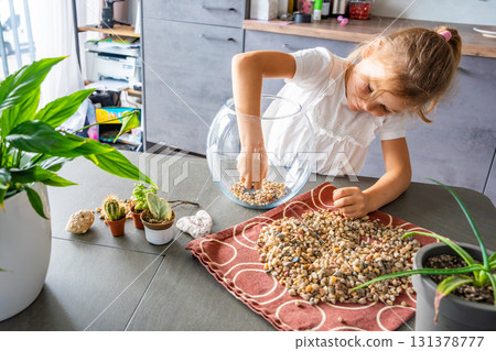 Little girl pouring stones into glass container to create drainage for future florarium or closed ecosystem. Hands-on learning, eco-friendly creativity and early involvement in nature-based projects. 131378777