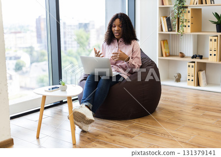 Young adult woman sitting on beanbag engaging in video call session using laptop in bright, well-lit interior with organized bookshelves and wooden floor, conveying productivity and comfort 131379141