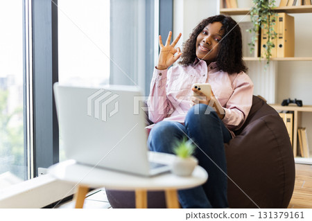 Young adult woman sitting on beanbag holding smartphone gesturing OK sign. Cozy home office environment with laptop, bright window, bookshelf. Concept of confidence work-life balance positivity 131379161