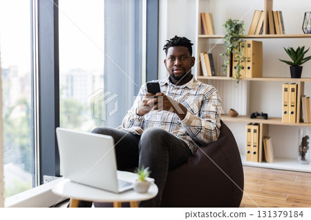 Young male adult using smartphone while sitting on beanbag, laptop on table nearby, indoors Young male adult using smartphone while sitting on beanbag, laptop on table nearby, indoors 131379184
