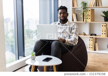 Young adult male sitting in a cozy room with laptop, working productively near window. 131379198