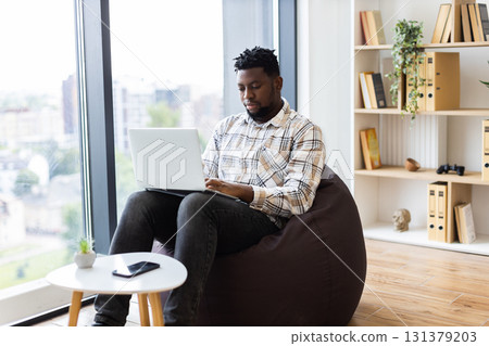 Young adult male focusing on laptop in bright casual interior with a window view Young adult male focusing on laptop in bright casual interior with a window view 131379203