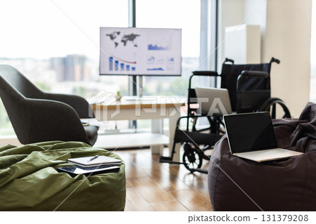 Bright office setup featuring wheelchairs, laptops on bean bags, large screen displaying graphs Bright office setup featuring wheelchairs, laptops on bean bags, large screen displaying graphs 131379208