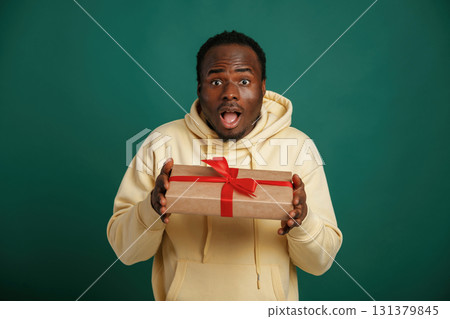 In shock. Holding gift box. African American man is standing against green background in the studio 131379845
