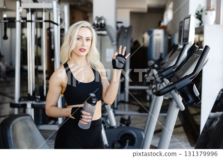 Athletic woman showing ok sign while holding water bottle in gym 131379996
