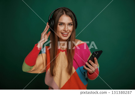 Moment of enjoying music, in headphones. Young woman is standing against green background in the studio Moment of enjoying music, in headphones. Young woman is standing against green background in the studio 131380287