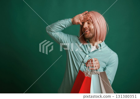 Hand near head. Holding shopping bags. Gay man with pink hair is standing against green background Hand near head. Holding shopping bags. Gay man with pink hair is standing against green background 131380351