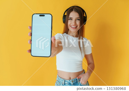 Headphones on head, holding smartphone with copy space. Young woman is against yellow background Headphones on head, holding smartphone with copy space. Young woman is against yellow background 131380968
