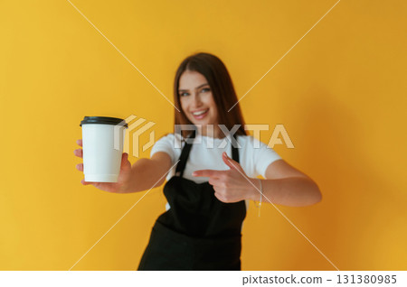 Black and white coffee worker uniform, holding cup of drink. Young woman is against yellow background Black and white coffee worker uniform, holding cup of drink. Young woman is against yellow background 131380985