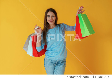 Happy facial expression, with shopping bags. Young woman is against yellow background 131381003