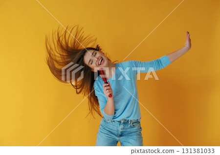 Singing into hairbrush. Young woman is against yellow background 131381033