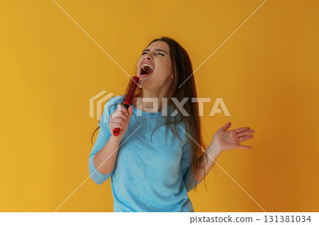 Singing into hairbrush. Young woman is against yellow background 131381034