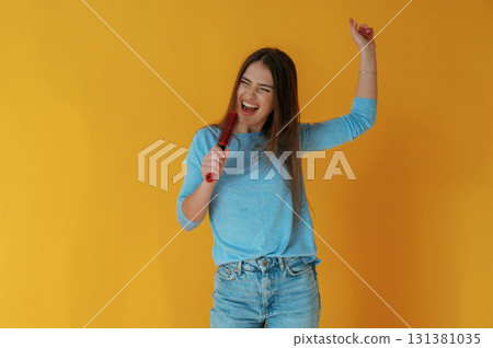 Singing into hairbrush. Young woman is against yellow background 131381035