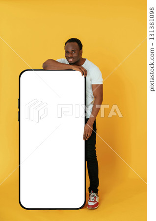 Leaning on the large smartphone for copy space. Black man is in the studio against yellow background 131381098