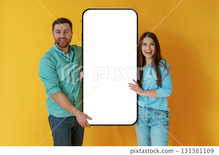 Display for copy space. Man and woman are standing with large smartphone against yellow background Display for copy space. Man and woman are standing with large smartphone against yellow background 131381109