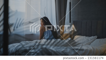 Woman in Nightwear Sits on Bed by Window in Soft Morning Light Woman in Nightwear Sits on Bed by Window in Soft Morning Light 131381174