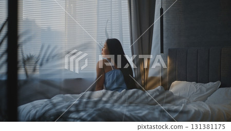 Woman in Nightwear Sits on Bed by Window in Soft Morning Light 131381175