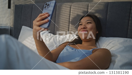 Young Woman Lies in Bed With Smile, Holding Smartphone During Video Call Young Woman Lies in Bed With Smile, Holding Smartphone During Video Call 131381190