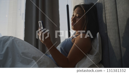 Beautiful Young Women Sitting in Bed and Scrolls Through News Feed on Smartphone Beautiful Young Women Sitting in Bed and Scrolls Through News Feed on Smartphone 131381332