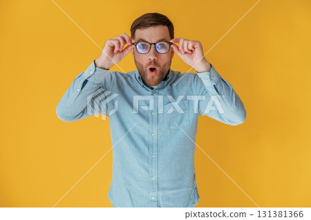 Shocked, touching glasses. Attractive man in blue shirt is against yellow background 131381366