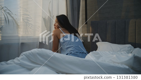Smiling Sleepily Woman, Sits Up in Bed By Sunny Window, Stretches Cozily Smiling Sleepily Woman, Sits Up in Bed By Sunny Window, Stretches Cozily 131381380