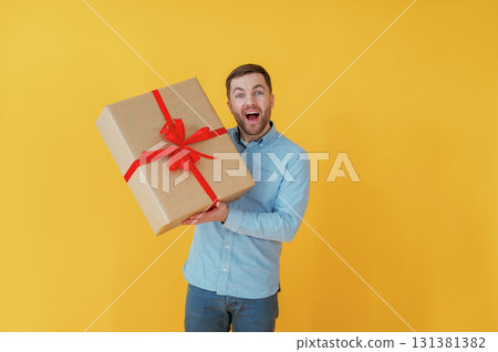 Surprised, holding gift box. Attractive man in blue shirt is against yellow background 131381382