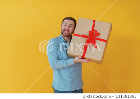 Surprised, holding gift box. Attractive man in blue shirt is against yellow background 131381383