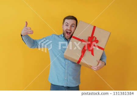 Surprised, holding gift box. Attractive man in blue shirt is against yellow background 131381384
